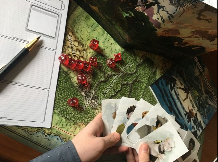 Person playing Dungeons and Dragons, focus on hands, board and cards with notepad and pen for scoring